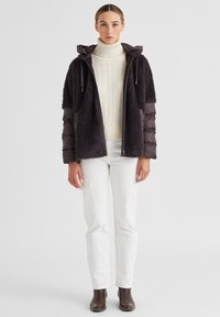 Dunkelbraune Kapuzenjacke aus strukturiertem Fleece mit gepolsterten Ärmeln, über einem cremefarbenen Rollkragenpullover getragen, kombiniert mit weißen Hosen und braunen Stiefeln.