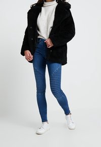 Černá umělá kožešinová bunda s kapucí, šedý pletený svetr, modré skinny džíny s žebrovanými detaily a bílé tenisky.