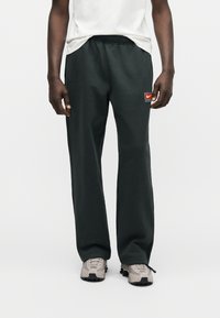 Άνδρας που φοράει σκούρα πράσινη φόρμα Nike με ελαστική μέση και άσπρα αθλητικά παπούτσια, στέκεται μπροστά σε ένα απλό φωτεινό φόντο.