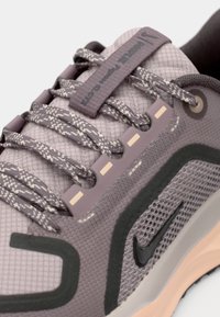 Gros plan d'une basket Nike grise avec des lacets à motifs, un tissu en mesh, un logo Swoosh noir et une semelle couleur pêche.