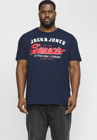 Camiseta de algodón azul marino con texto en rojo y blanco en negrita "JACK & JONES Superior International Standard" en el pecho, con diferentes tipografías.
