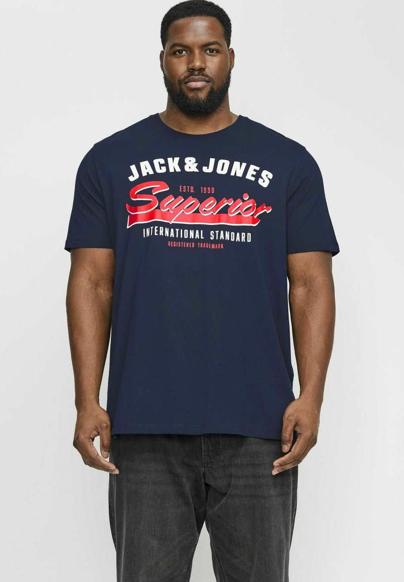 Tumši zils kokvilnas T-krekls ar izteiksmīgu sarkano un balto tekstu "JACK & JONES Superior International Standard" krūšu daļā dažādos fontu stilā.
