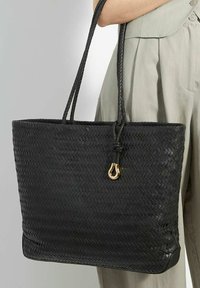 Black woven tote bag with thick, braided handles and a gold accent. Boxy shape with a textured finish and a flat base.