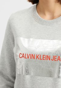 Sudadera gris con escote redondo, que presenta un panel de textura plateada agrietada y el texto rojo "CALVIN KLEIN JEANS" en la parte frontal.