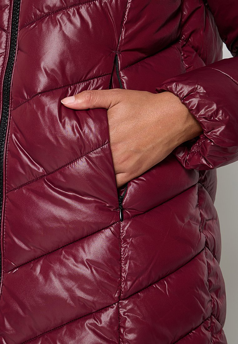 Hand gedeeltelijk gestoken in de ritszak aan de zijkant van een bordeauxrode gewatteerde pufferjack met een elastische manchette.