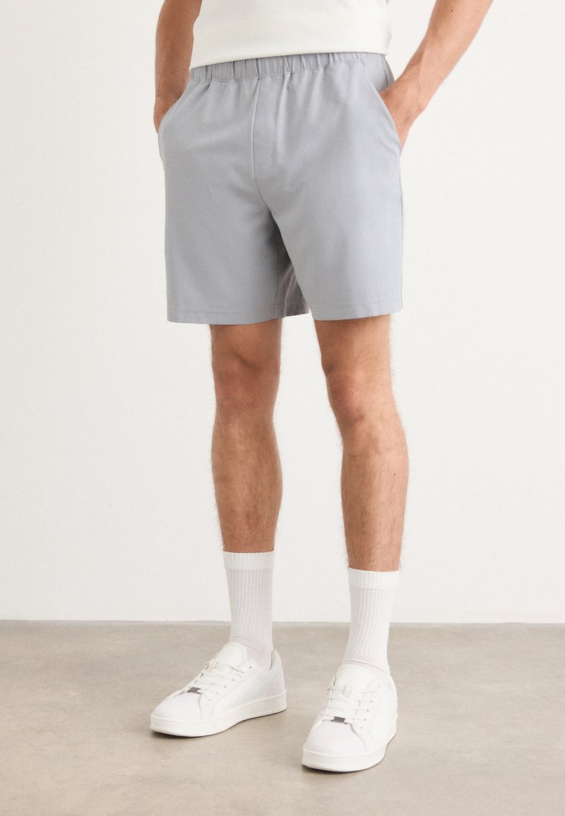 Grå shorts med elastisk linning, sidelommer og en avslappet passform, kombinert med hvite joggesko og knelange hvite sokker.