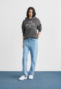 Γυναίκα όρθια, φορώντας γκρι φούτερ New Balance με κουκούλα, ανοιχτό μπλε τζιν με ρίγες και λευκά αθλητικά παπούτσια σε μπλε χαλί.