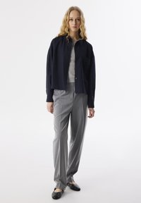 Marineblauw overhemd, grijze trui, grijze op maat gemaakte broek en zwarte platte schoenen met studs. Zachte texturen en een ontspannen pasvorm.