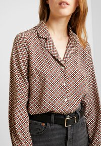 Camicia a maniche lunghe con bottoni in un motivo floreale con accenti rossi e blu navy su sfondo crema; abbinata a jeans scuri a vita alta e una cintura nera.