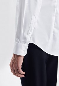 Witte dress-shirt met knoopmanchetten, glad geweven stof, gebogen zoom, gecombineerd met zwarte, aansluitende broek. Hand rustend op de dij.