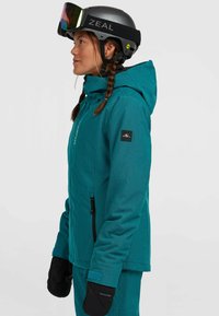Teal sneeuwjack met een gestructureerde afwerking, verstelbare capuchon, ritssluitingen en zwarte handschoenen. Draag met een grijze helm en kleurrijke goggles.