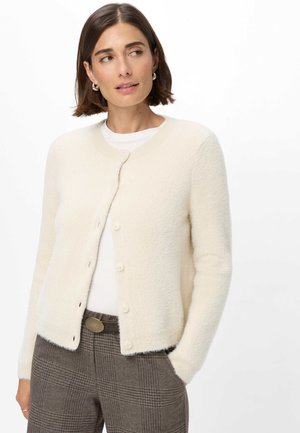 Femme aux cheveux courts et bruns porte un cardigan blanc boutonné par-dessus un haut blanc et un pantalon marron à carreaux, debout avec une main dans la poche.