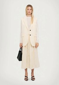 Nainen, joka on pukeutunut pitkään kermaiseen liiviin ja leveisiin housuihin, pitää mustaa käsilaukkua ja seisoo vaaleaa taustaa vasten.