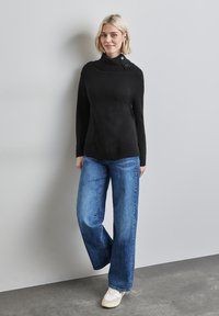 Pull en tricot noir avec un col montant et des détails de boutons, associé à un pantalon large en jean bleu clair et des baskets blanches sur un fond gris.
