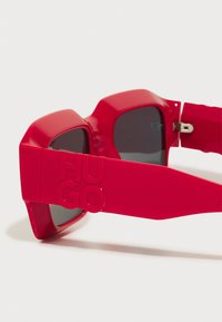 Red sunglasses with a chunky, angular shape. Made of glossy plastic, featuring dark lenses and raised "HUGO" logo on the temple.
