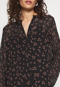 Kvinde iført en gennemsigtig sort bluse med små rosa blomsterprint, knaplukket foran, med krøllet brunt hår synligt.