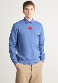 Svetlomodrý sweatshirt s rebrovanými manžetami a lemom, s červeným obdĺžnikovým logom. Pod ním je viditeľná svetlomodrá košeľa s golierom.