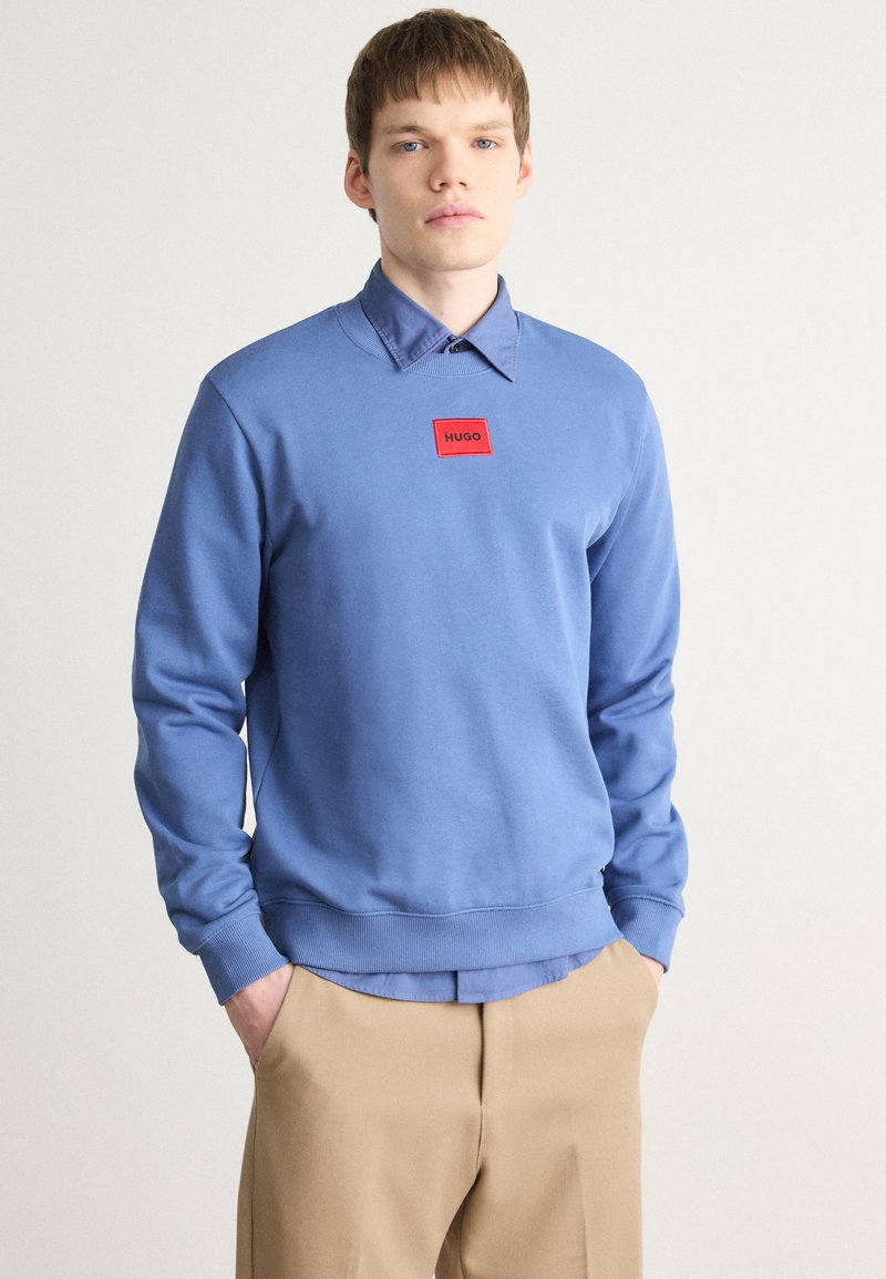 Svetlomodrý sweatshirt s rebrovanými manžetami a lemom, s červeným obdĺžnikovým logom. Pod ním je viditeľná svetlomodrá košeľa s golierom.