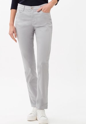 Vrouw draagt lichtgrijze rechte broek, zwart shirt en witte sneakers, met één hand in de zak tegen een witte achtergrond.