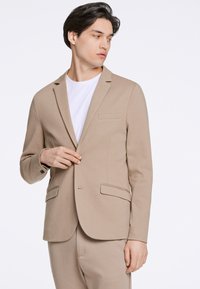 Beżowa marynarka o dopasowanym kroju, z zapięciem na jeden guzik, klapami typu notch oraz dwoma przednimi kieszeniami. Materiał wydaje się gładki i teksturowany.