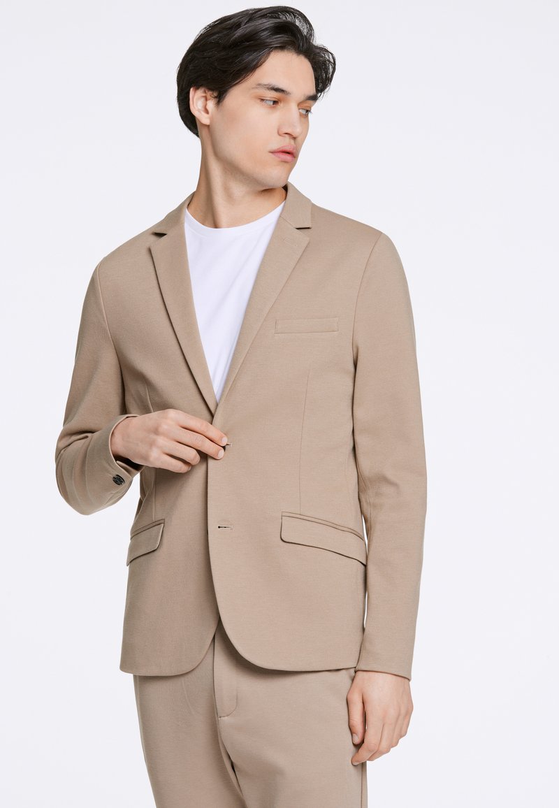 Beżowa marynarka o dopasowanym kroju, z zapięciem na jeden guzik, klapami typu notch oraz dwoma przednimi kieszeniami. Materiał wydaje się gładki i teksturowany.