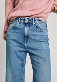 Vaqueros de denim azul claro con cintura alta, bolsillos delanteros y costuras en contraste. Textura suave, corte recto, combinados con un suéter de punto rosa.