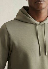 Olijfgroene hoodie van zacht materiaal, met een trekkoordcapuchon met metalen aglets en een strak, minimalistisch ontwerp.
