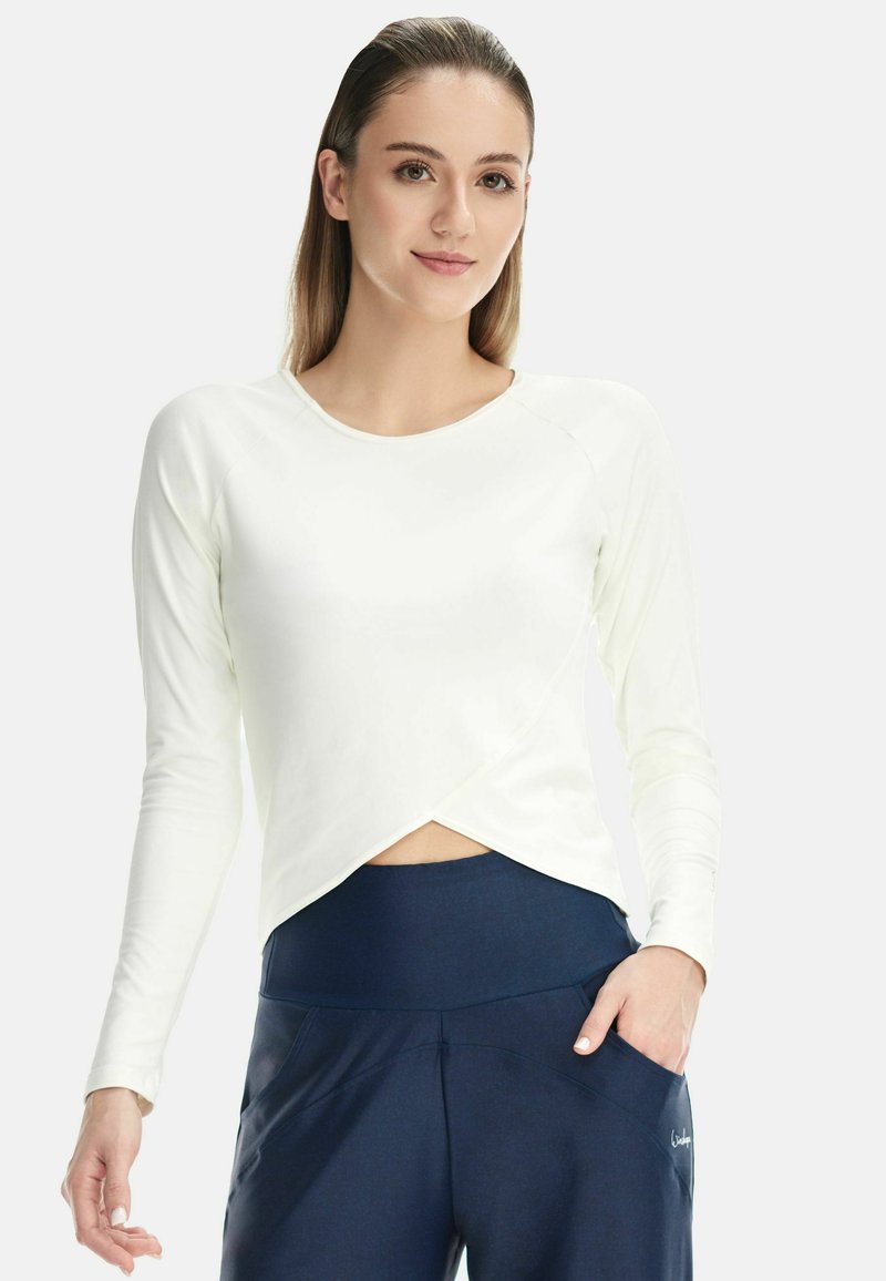 Long-sleeve top in light cream, featuring a wrap design. Worn with dark blue high-waisted pants with side pockets and a textured finish.