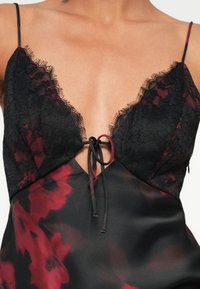 Abito slip in satin rosso floreale con pizzo nero, scollatura profonda e spalline regolabili, caratterizzato da un dettaglio centrale con nodo.