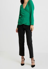 Grüne Wickelbluse mit langen Ärmeln und strukturiertem Stoff, mit gebundenem Taillenakzent. Kombiniert mit schwarzen, verkürzten Jeans und schwarzen, geschnürten High Heels.