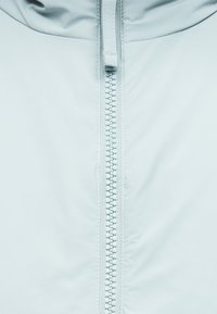 Κοντινή λήψη ενός λευκού φερμουάρ μπουφάν στο κέντρο κάθετα, που δείχνει τα δόντια του φερμουάρ και τον μοχλό του πάνω σε λείο ύφασμα.