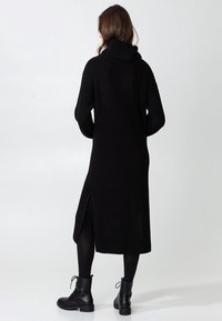 Abito nero a maglia con colletto alto, maniche lunghe, spacchi laterali e silhouette rilassata. Indossato con stivali neri alla caviglia e collant.