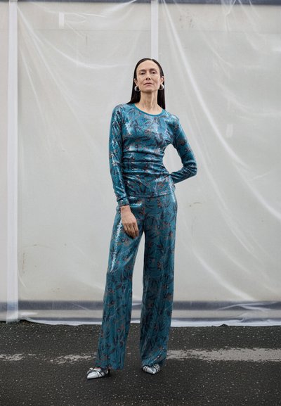 Haut métallique bleu sarcelle à manches longues avec motif floral, assorti à un pantalon à jambes larges. Le mannequin porte des chaussures rayées à bout pointu et des boucles d'oreilles créoles.