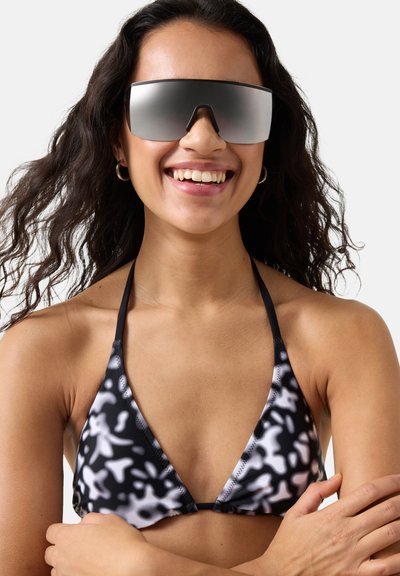 Frau mit langen lockigen Haaren, die ein schwarz-weiß gemustertes Bikinioberteil und große, reflektierende Sonnenbrillen trägt, lächelt mit verschränkten Armen.