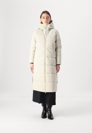 Gevoerde lange jas in lichtbeige met een capuchon, voorzien van een rits aan de voorkant en zijzakken, gecombineerd met een zwarte jurk en zwart kniehoge laarzen.