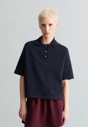 Person mit kurzem blondem Haar, die ein lockeres marineblaues Kurzarmhemd und einen kastanienbraunen Rock trägt und vor einem schlichten hellgrauen Hintergrund steht.