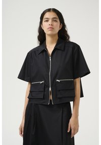 Černá cropped bunda s předním zipem, krátkými rukávy, dvěma kapsami na zip a plochými kapsami na dně, vyrobená z hladkého materiálu.