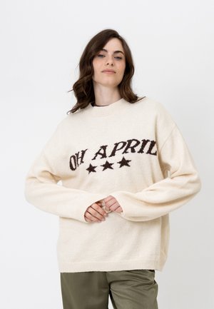 Jeune femme aux cheveux bruns ondulés porte un pull crème avec l'inscription "OH APRIL" et trois étoiles, associé à un pantalon vert olive sur fond blanc.
