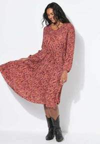 Femme portant une robe à manches longues, à motifs orange et violet avec une taille froncée, accompagnée de bottes de cow-boy noires, se tenant devant un mur blanc.