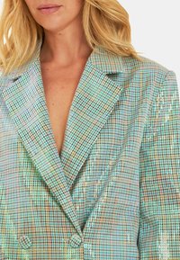 Blazer doppiopetto in tessuto a quadri metallici, con effetto scintillante, rever a scollo e due bottoni frontali. Toni di blu chiaro, verde e beige.