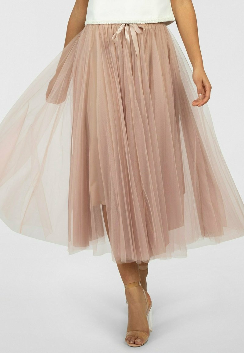 Femme portant une jupe midi en tulle fluide rose clair avec une ceinture nouée à la taille et des sandales à talons hauts beiges, sur un fond uni.