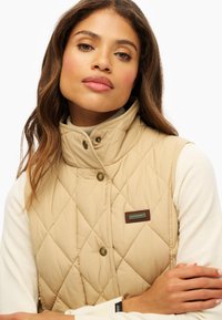 Gilet matelassé beige avec col montant et boutons-pression. Présente un écusson logo marron. Porté par-dessus un haut à manches longues de couleur claire.