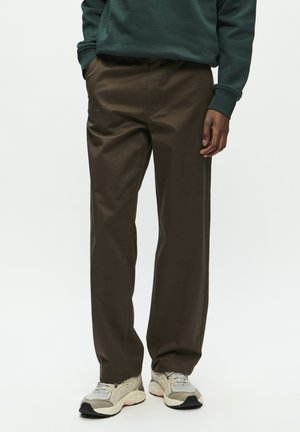 Uomo che indossa pantaloni marrone scuro a gamba dritta, felpa verde e scarpe da ginnastica grigio chiaro, in piedi con le mani nelle tasche.