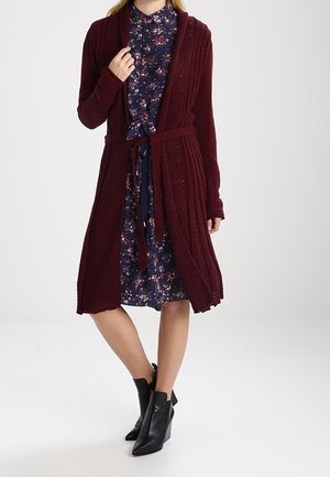 Femme portant un cardigan en maille bordeaux ceinturé sur une robe fleurie bleu marine avec des bottines noires, debout devant un fond uni.