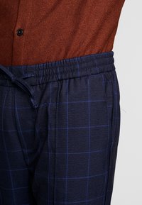 Närbild av marinblå rutiga byxor med elastisk midja och dragsko, i kombination med en rostfärgad skjorta med knappar.