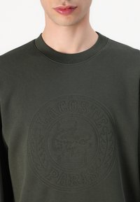 Sweat-shirt vert foncé à encolure ronde, présentant un logo en relief texturé au centre, avec des poignets et un ourlet côtelés.