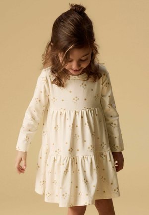 Jeune fille regardant vers le bas, portant une robe à manches longues crème avec de petites impressions florales jaunes sur un fond beige uni.