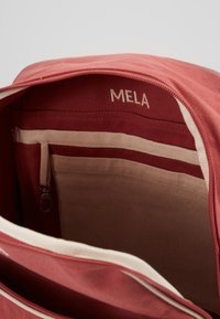 Sarkanas auduma somas iekšpuse ar gaiši pelēku oderi, kurā ir rāvējslēdzēja kabata un etiķete ar uzrakstu "MELA." Gluda auduma tekstūra.