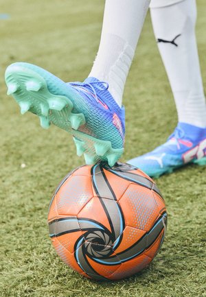 Puma-Fußballschuh mit einem blauen Farbverlauf-Design und grünen Stollen, der auf einem orange-schwarz gemusterten Fußball auf Gras drückt.