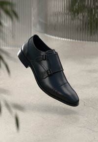 Sort læder double monk strap dress sko svævende over let revnet gulv med slørede planter og bølget væg i baggrunden.